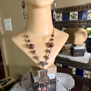 Puede incluir: Un conjunto de collar y pendientes de estilo vintage, exhibido en un busto de maniquí. El collar presenta un colgante con una figura femenina, cuentas rosas y perladas, y detalles rojos. Los pendientes tienen un diseño similar con cuentas en forma de lágrima.