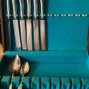 May include: A vintage silverware set in a teal felt-lined wooden case. The set includes knives and spoons with ornate silver handles. The case is divided into compartments to hold the utensils. The silverware is arranged neatly, suggesting a formal dining setting.