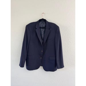 May include: A navy blue blazer with two buttons and two front pockets hangs on a black hanger. The blazer has a notched lapel and a single vent in the back. The jacket is made of a smooth, solid fabric.