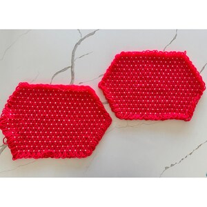 May include: Two red, hexagonal crochet potholders. The potholders are made of a tightly woven red yarn with a scalloped edge. The potholders are displayed on a white marble surface.