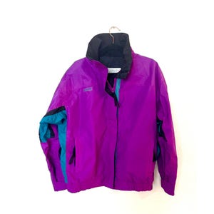 Könnte beinhalten: Eine vintage lila und türkis Columbia Jacke mit schwarzem Kragen und Akzenten. Die Jacke hängt an einem goldenen Kleiderbügel. Die Jacke hat einen Reißverschluss und ein kleines Logo auf der Brust.