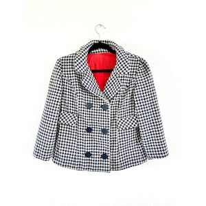 May include: A black and white houndstooth double-breasted jacket with navy blue buttons and a red lining. The jacket has two front pockets and is hanging on a black hanger against a white background.