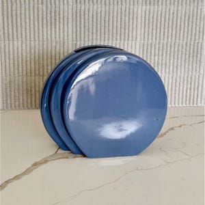 May include: A blue ceramic vase with a glossy finish. The vase has a unique design with curved, layered sections. The vase is a solid blue color and sits on a marble-like surface. The background is a textured, light-colored wall.