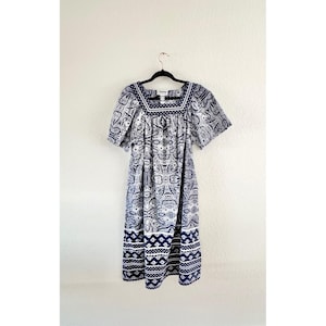 May include: A blue and white paisley print dress with a square neckline and short sleeves. The dress is hanging on a black hanger against a white wall. The dress has a white and blue border at the neckline and hem.