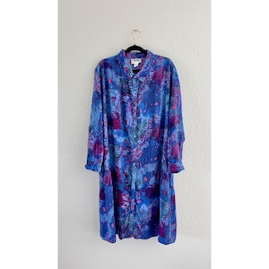 May include: A long-sleeved dress in shades of blue and purple, featuring a botanical pattern. The dress has a collared neckline and button-down front. The dress is hanging on a black hanger against a white wall.