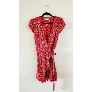 May include: A red wrap dress featuring a repeating cream-colored abstract spot pattern, short sleeves, and a ruffled hemline. The garment includes a tie closure at the waist and a label at the neckline that reads XII PALMS.