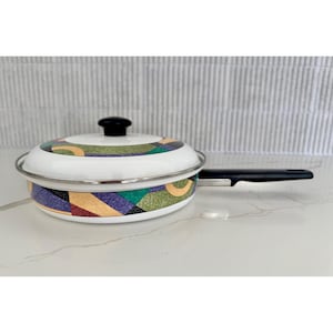 May include: A white enamel pan with a black handle and lid. The pan features a colorful geometric pattern in shades of purple, green, yellow, and red. The pan is approximately 8 inches in diameter.