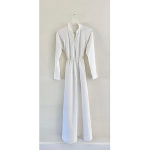 May include: A white dress with long sleeves and a zipper closure. The dress has a high neckline and a fitted waist, with a flowing skirt. The dress is hanging on a white hanger against a white wall.