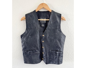 Op de afbeelding: Een zwart leren vest met een V-hals, vijf goudkleurige knopen aan de voorzijde en twee zakken onderaan. Het binnenlabel vermeldt Genuine Leather. Het kledingstuk hangt aan een houten kledinghanger tegen een witte muur.
