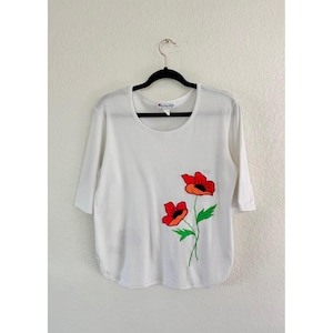 May include: White scoop-neck shirt with three-quarter sleeves. The shirt features an embroidered design of two red poppies with orange centers and green stems and leaves. The shirt is hanging on a black hanger against a white wall.