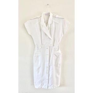 May include: A white short-sleeved dress featuring a collared neckline, shoulder epaulets, and a button-down front closure. The garment includes two front hip pockets and a cinched waist design, presented on a hanger against a plain background.