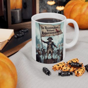 May include: White ceramic mug with a Halloween-themed illustration. The design features a skeletal figure in colonial attire holding a banner that reads "No Reanimation Without Representation!" surrounded by other skeletal figures. The mug is filled with dark liquid.
