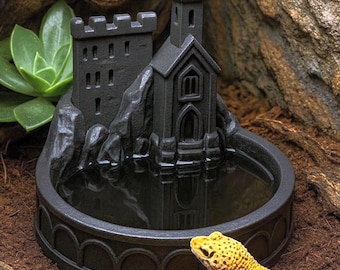 May include: A dark gray, circular water dish featuring a miniature castle structure with a tall tower and crenelated walls. The basin holds water, and the exterior base is decorated with a series of arched carvings along the outer rim.
