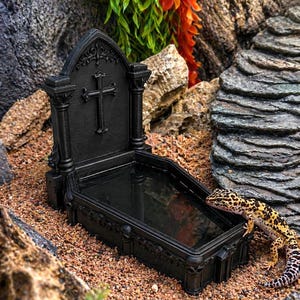 May include: A black, coffin-shaped water dish for reptiles featuring a raised headstone with a cross emblem and decorative skull accents along the base. A yellow and black spotted leopard gecko rests beside the dark, rectangular basin.