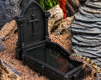 May include: Black gothic-style reptile water dish with a cross design. The dish is shaped like a coffin and is filled with water. A leopard gecko is perched on the edge of the dish. The dish is surrounded by rocks and plants.