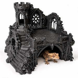 May include: A black, three-dimensional printed reptile hide shaped like a ruined gothic castle with a tower, stone stairs, and arched windows. A brown and tan crested gecko is positioned underneath the main archway of the structure.