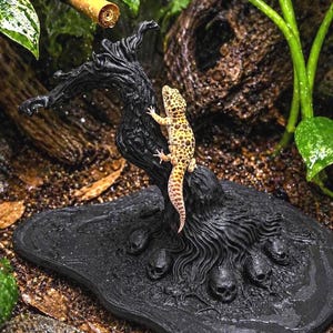 May include: A detailed shot of a reptile habitat decoration. A yellow and brown leopard gecko climbs a black, textured tree-like structure. The base features skull details. The background includes greenery and a bamboo-like structure.