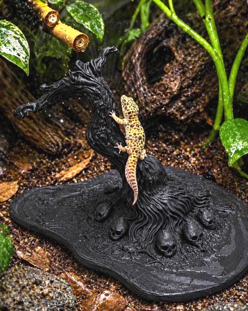 May include: A detailed shot of a reptile habitat decoration. A yellow and brown leopard gecko climbs a black, textured tree-like structure. The base features skull details. The background includes greenery and a bamboo-like structure.
