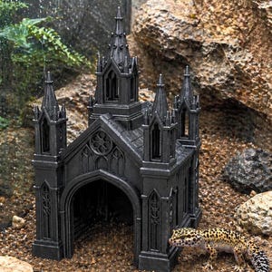 Puede incluir: Decoraci&oacute;n de acuario negra de estilo g&oacute;tico con forma de catedral, con agujas y entradas arqueadas. Un gecko leopardo con manchas amarillas y negras est&aacute; cerca de la estructura. El adorno est&aacute; colocado sobre un lecho de peque&ntilde;os guijarros.