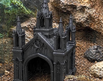 May include: A black gothic-style aquarium decoration shaped like a cathedral, featuring spires and arched doorways. A leopard gecko with yellow and black spots is near the structure. The decoration is placed on a bed of small pebbles.