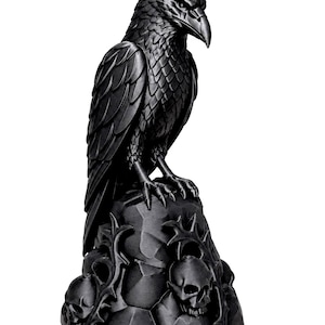 Peut inclure: Une sculpture noir mat représentant un corbeau perché sur une formation rocheuse dentelée. La base du rocher est ornée de trois crânes humains et de structures épineuses ressemblant à des vignes. Le corbeau a des plumes détaillées.
