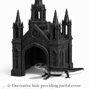 Puede incluir: Escondite decorativo negro de estilo g&oacute;tico para reptiles, que se asemeja a una iglesia en miniatura con agujas y puertas arqueadas. Un lagarto blanco y negro est&aacute; posicionado frente al escondite. El texto de la imagen dice: "Escondite decorativo que proporciona cobertura parcial con un estilo g&oacute;tico oscuro."