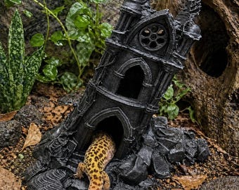Puede incluir: Una torre de estilo gótico en color negro con una ventana circular y aberturas arqueadas, que sirve como escondite para reptiles. Un gecko leopardo con manchas amarillas y marrones está parcialmente dentro de la entrada arqueada inferior.
