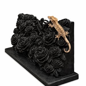 May include: A rectangular black base supports a vertical panel featuring a dense, sculpted arrangement of black roses. A light tan crested gecko with a patterned back and tail is positioned on the right side of the floral relief.