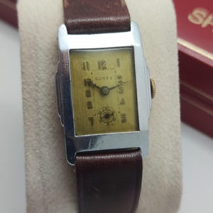May include: A vintage rectangular watch with a silver-tone case and a brown leather band. The watch face is gold-toned with black numerals and the brand name "CONTY". The watch is displayed on a white surface.