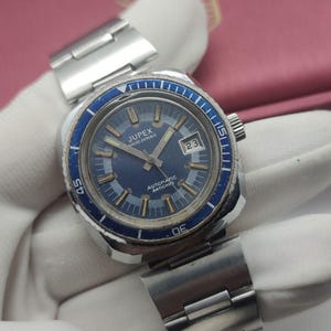Può includere: Un orologio da polso automatico Jupex con quadrante rotondo blu, lancette color argento e indici delle ore. L'orologio presenta una lunetta girevole blu con numeri bianchi, una finestrella della data a ore tre e un cinturino a maglie in acciaio.