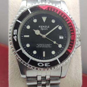 Può includere: Un orologio da polso Kienzle Sport con quadrante nero, lancette color argento e indici circolari. La lunetta presenta una combinazione di colori nero e rosso con numeri bianchi. È dotato di un cinturino a maglie in acciaio inossidabile.