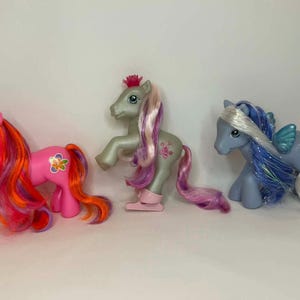 May include: Three toy ponies in a row. The first is pink with a flower and long, colorful hair. The second is silver with pink hair and a crown. The third is blue with wings and white hair.