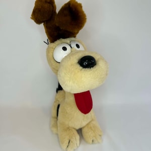 May include: A soft toy dog with a cream-coloured body, large white eyes, and brown ears. The dog has a black nose, a red tongue, and black spots on its back. The toy is sitting upright on a white surface.