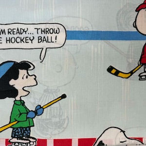 Puede incluir: Tela azul claro con los personajes de Peanuts, Lucy y Charlie Brown, jugando al hockey. Lucy lleva una camisa verde y un gorro azul, sosteniendo un palo de hockey. Charlie Brown lleva una chaqueta y un gorro rojos. El texto dice: "OKAY, I'M READY... THROW ME THE HOCKEY BALL!"