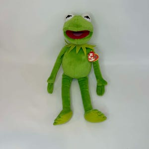 以下が含まれることがあります： 緑色のカーミット・ザ・フロッグのぬいぐるみ。赤い口、白い目、黄色の襟が特徴です。おもちゃには赤いハート型のタグが付いています。カエルは白い背景を背景に立っています。