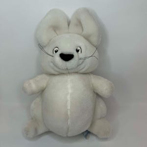 Peut inclure: Une peluche blanche en forme de lapin avec de grandes oreilles tombantes, des yeux noirs et un nez noir. Le lapin a un sourire cousu et de longues moustaches. Le jouet en peluche est fait d'une matière duveteuse.