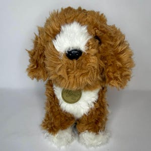 May include: A soft toy dog with brown and white fur. The toy features a white patch on its face and chest, with brown fur covering the rest of its body. It has a black nose and a tag that reads "Miyoni".