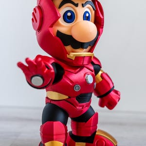 Pu&ograve; includere: Una statuina rossa e oro di Mario in un'armatura di Iron Man. La figura ha un elmo rosso con dettagli dorati, baffi neri e occhi blu. L'armatura ha un aspetto metallico, con dettagli come rivetti e pannelli.