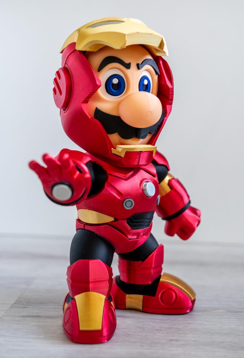 Pu&ograve; includere: Una statuina rossa e oro di Mario in un'armatura di Iron Man. La figura ha un elmo rosso con dettagli dorati, baffi neri e occhi blu. L'armatura ha un aspetto metallico, con dettagli come rivetti e pannelli.