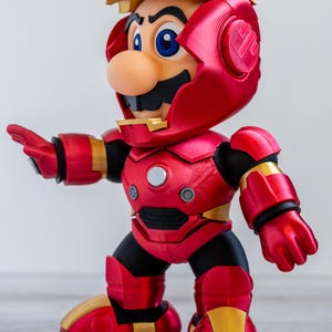 Pu&ograve; includere: Una statuina di Mario in un'armatura di Iron Man. La figura &egrave; principalmente rossa e dorata, con dettagli neri. L'elmo &egrave; parzialmente aperto, rivelando il volto di Mario. La figura &egrave; in posa con un braccio teso.