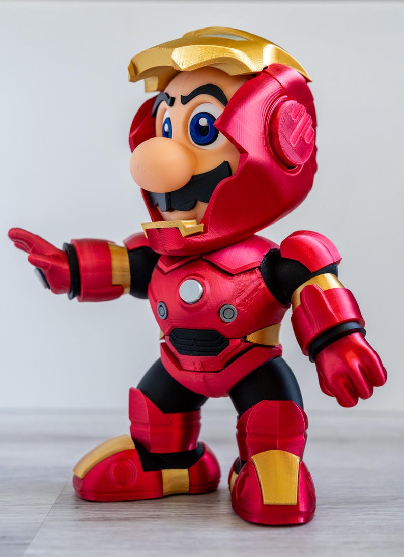 Pu&ograve; includere: Una statuina di Mario in un'armatura di Iron Man. La figura &egrave; principalmente rossa e dorata, con dettagli neri. L'elmo &egrave; parzialmente aperto, rivelando il volto di Mario. La figura &egrave; in posa con un braccio teso.