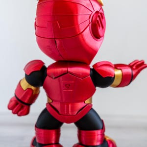 Pu&ograve; includere: Statuetta rossa e oro di un supereroe, vista da dietro. La figura ha una testa e un corpo arrotondati, con braccia e gambe articolate. Il design presenta una finitura metallica, con accenti dorati sul casco, sulle spalle e sulle articolazioni.