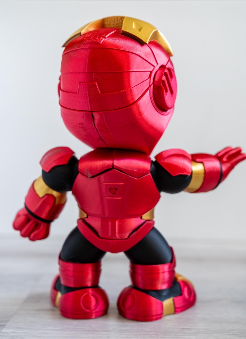 Pu&ograve; includere: Statuetta rossa e oro di un supereroe, vista da dietro. La figura ha una testa e un corpo arrotondati, con braccia e gambe articolate. Il design presenta una finitura metallica, con accenti dorati sul casco, sulle spalle e sulle articolazioni.
