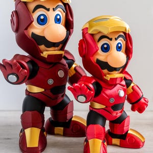 Pu&ograve; includere: Due statuette di Mario con l'armatura di Iron Man. Le figure sono principalmente rosse con dettagli dorati, con l'iconico baffo di Mario e occhi blu. L'armatura ha un design meccanico dettagliato.