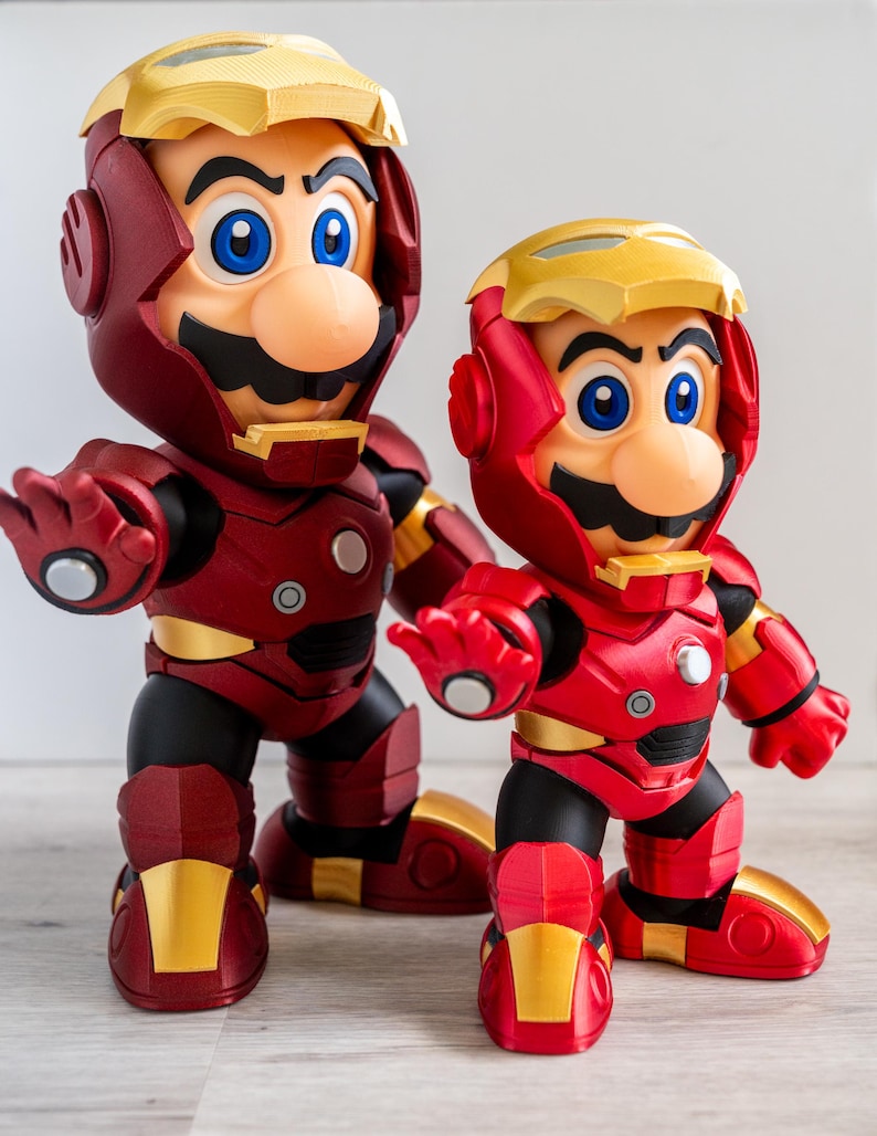 Pu&ograve; includere: Due statuette di Mario con l'armatura di Iron Man. Le figure sono principalmente rosse con dettagli dorati, con l'iconico baffo di Mario e occhi blu. L'armatura ha un design meccanico dettagliato.