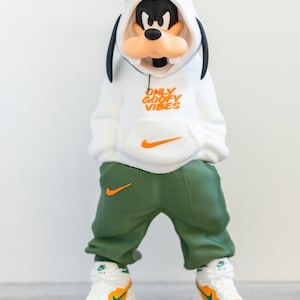May include: A figurine of Goofy wearing a white hoodie with "ONLY GOOFY VIBES" text and an orange Nike swoosh. He's also wearing green sweatpants with an orange Nike swoosh and white and orange sneakers. The figurine has a green hat.