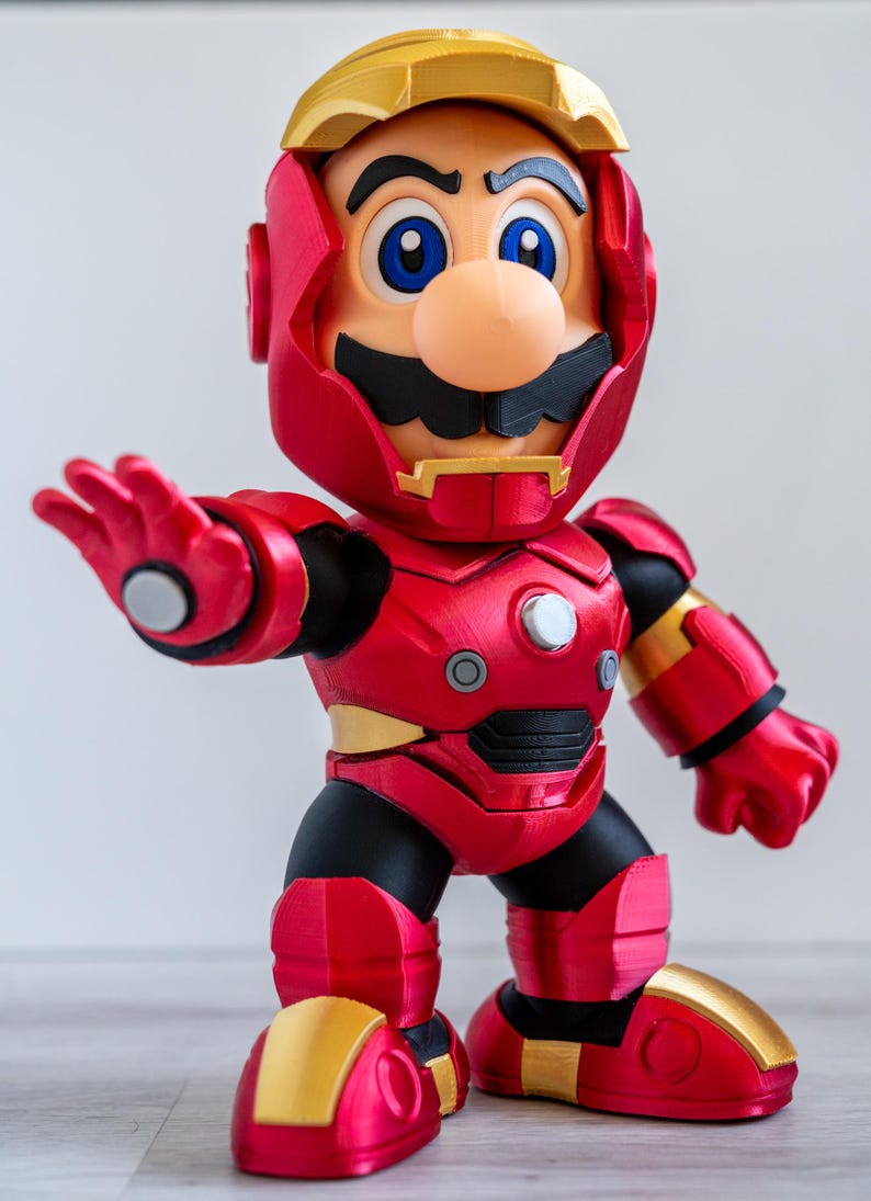 Pu&ograve; includere: Una action figure rossa e oro di un personaggio che &egrave; un mix tra Mario e Iron Man. La figura ha un'armatura rossa con dettagli dorati, un elmo dorato e la faccia di Mario. La figura &egrave; in posa con un braccio teso.