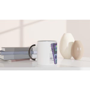 Op de afbeelding: Witte keramische mok met een zwart interieur en handvat. De mok heeft een paarse en groene afbeelding van een draagbare gameconsole. Het ontwerp is geplaatst tegen een rasterachtergrond.