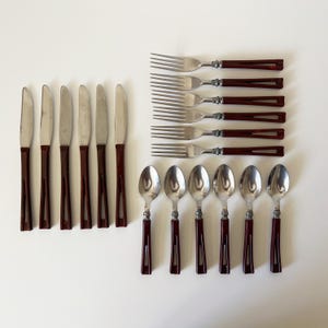May include: A set of vintage silverware with stainless steel blades and tines, and dark brown handles. The set includes six knives, six forks, and six spoons, arranged on a white surface. The silverware is likely from the mid-century.