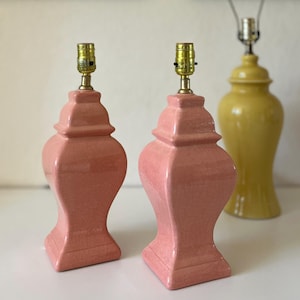 May include: Two matching table lamp bases with a glossy pink crackle glaze finish, featuring a square base and a tiered, urn-shaped body. Each lamp base is topped with a metallic gold-toned socket. A single yellow lamp base is visible behind.